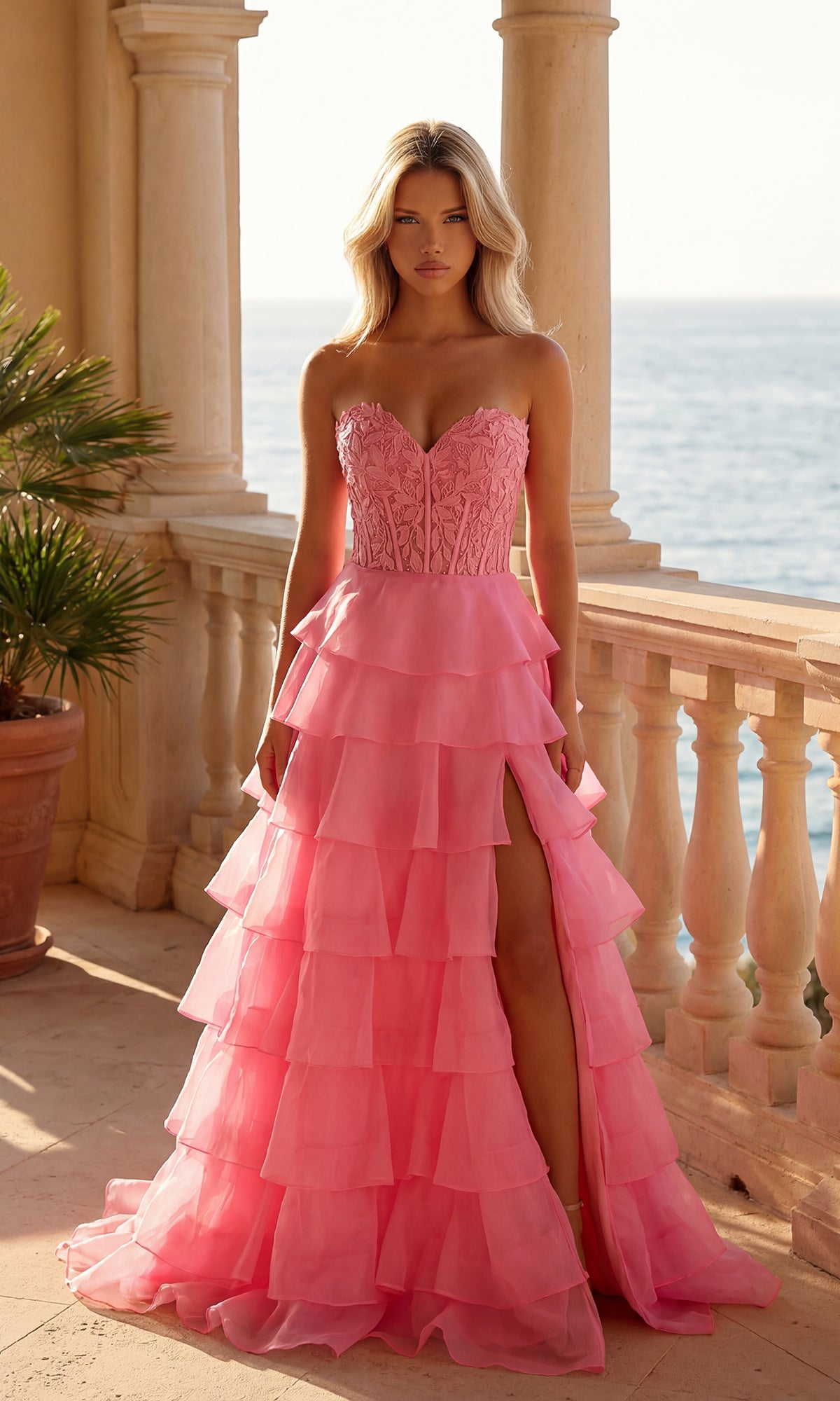 A woman in a bright pink Strapless Long Ruffled Prom Gown (PromGirl 250001) with a sheer corset-style bodice and thigh-high slit stands on a sunlit terrace overlooking the ocean.